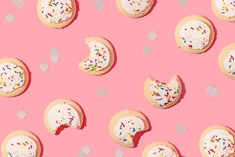 white frosted sprinkle sugar cookies with bites taken out of them, on a pink background with gold confetti.